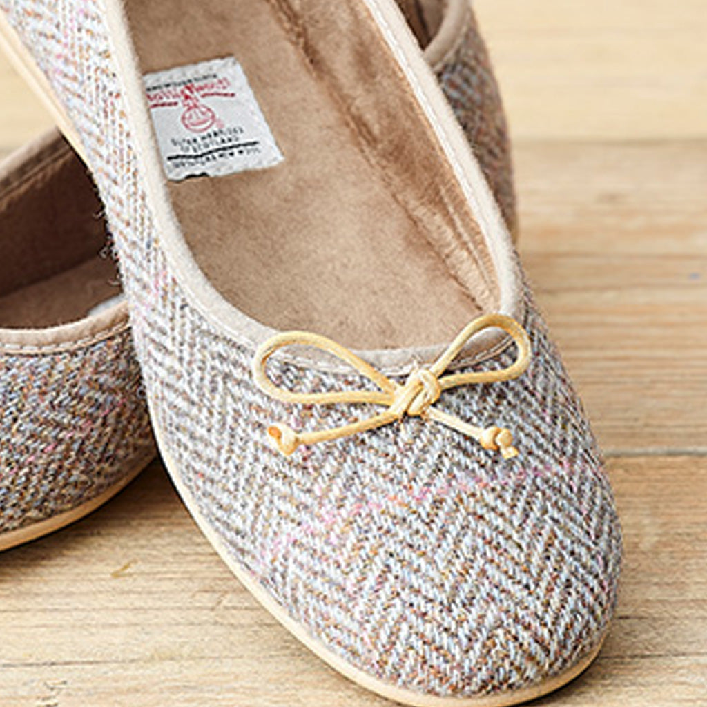 A close-up of a pair of women's herringbone slip-on flats in a beige, brown, and gray woven pattern, featuring a light tan leather bow on the front. The shoes are displayed on a light wooden surface.