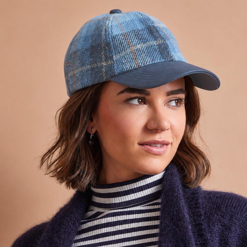 A woman wearing a blue plaid baseball cap with a navy blue visor, styled with a striped turtleneck and a dark knit cardigan against a solid beige background.