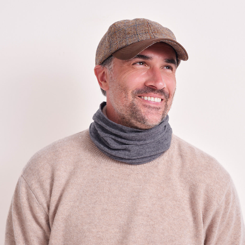 A smiling man wearing a brown tweed flat cap, a dark gray knit neck warmer, and a beige knit sweater, photographed against a plain light background.