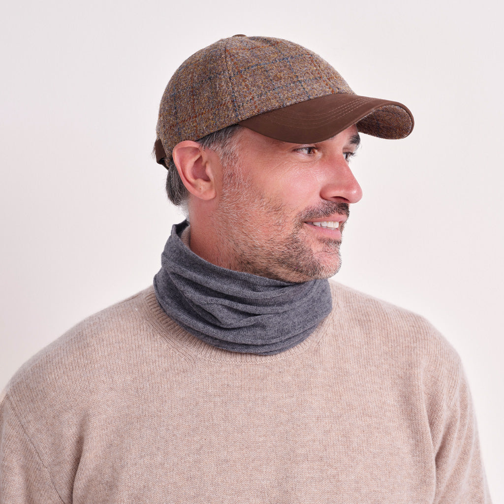 A man wearing a brown tweed baseball cap, a dark gray knit neck warmer, and a beige crewneck sweater, shown in profile against a plain light background.