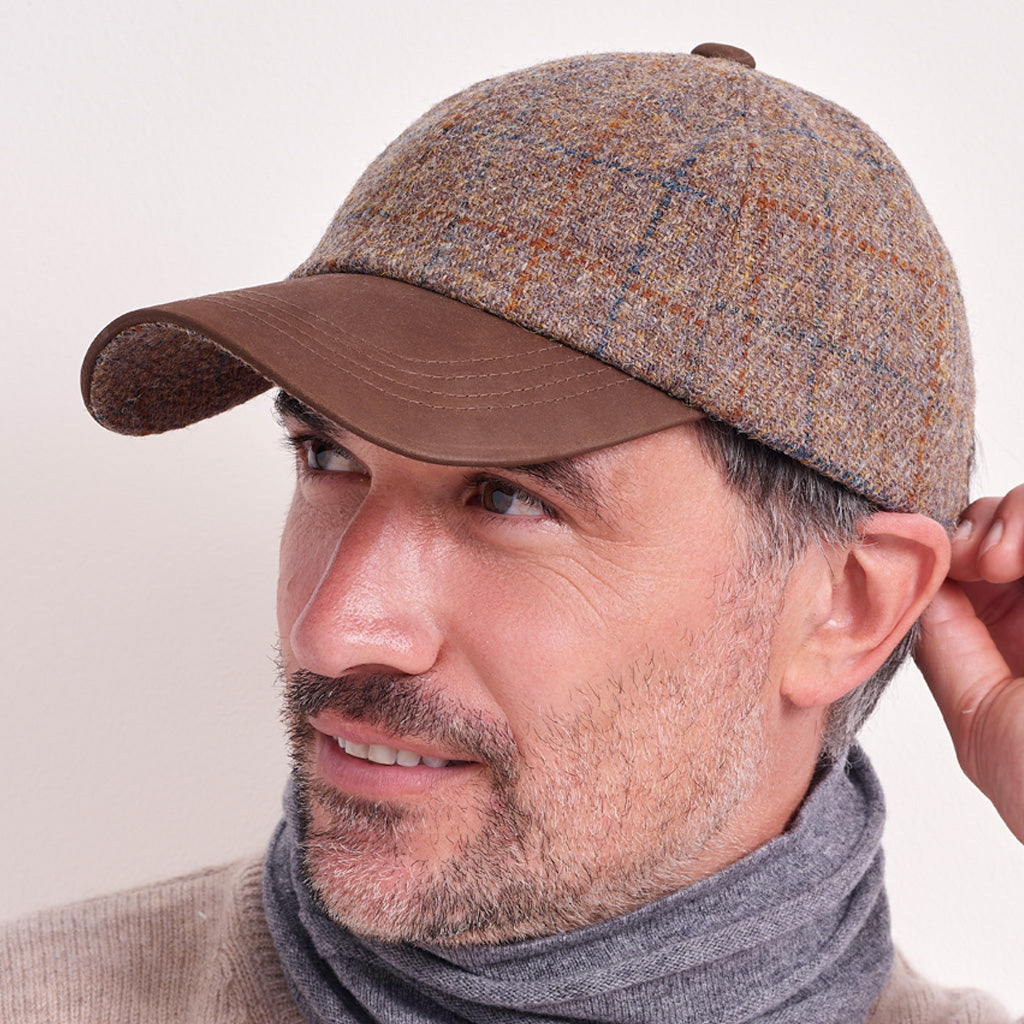 A man wearing a brown tweed baseball cap with a solid brown visor, paired with a gray knit neck warmer and beige sweater, against a plain light background.