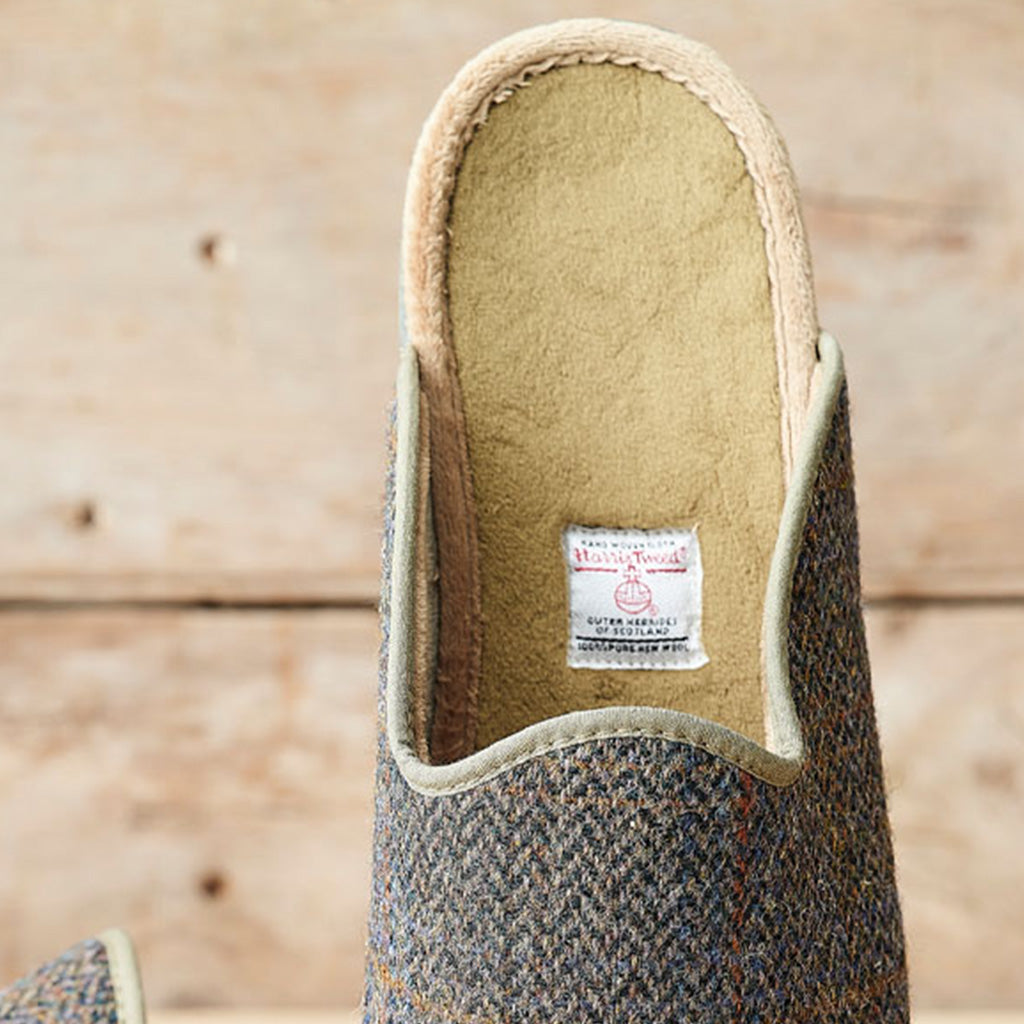 A close-up of the inside of a Tweed Mill slouch slipper, showing the beige fleece lining and the brand label, set against a rustic wooden background.