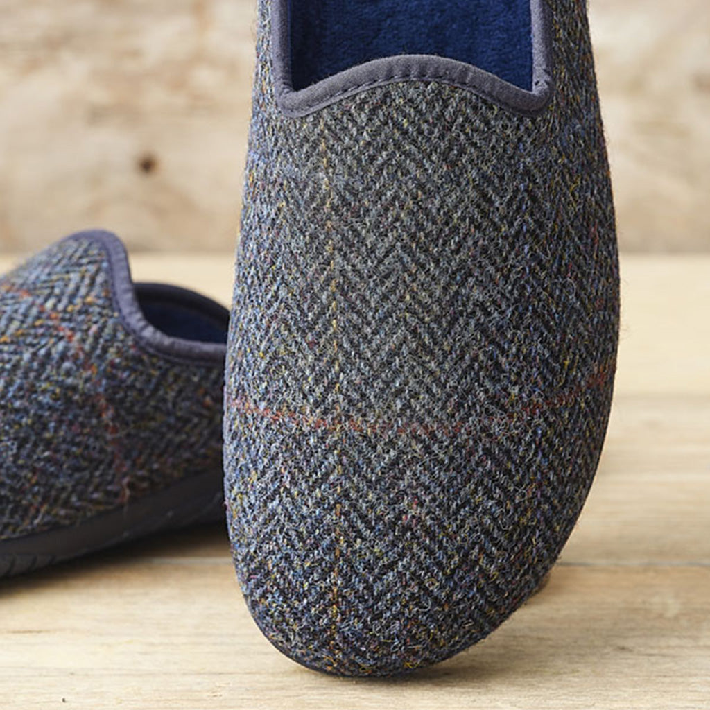 A close-up of a slip-on herringbone slipper in dark gray and blue tweed with a navy blue interior, shown on a light wood surface with another slipper blurred in the background.
