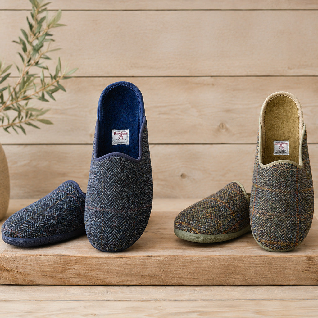 Three slip-on woven slippers displayed on a wooden surface against a light wood plank background. The left slipper is dark navy blue with a herringbone pattern and a blue interior.