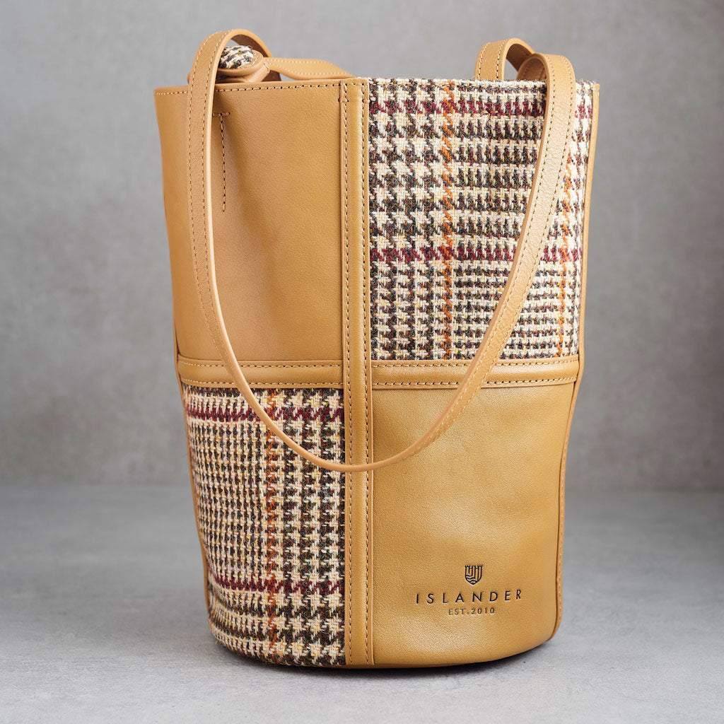 A tan leather bucket bag with a houndstooth patterned panel on the front, featuring a single shoulder strap and the ISLANDER logo embossed on the lower right front.