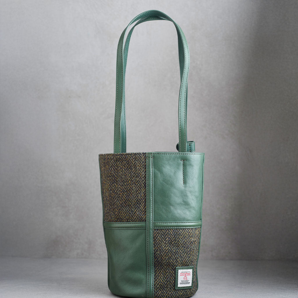 A green leather and herringbone tweed bucket bag with dual shoulder straps and a visible Woolworths label patch on the lower front, photographed against a neutral gray background.