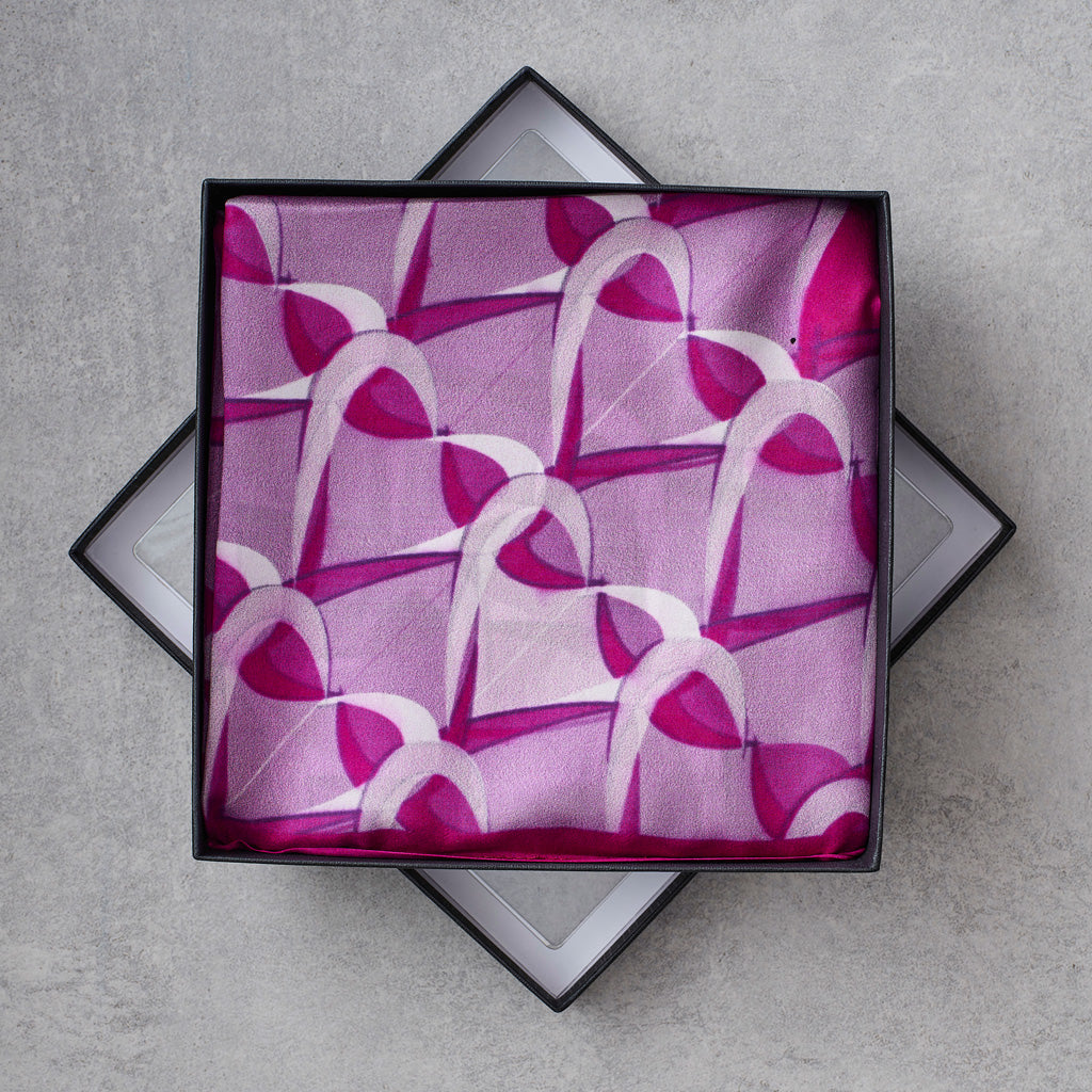A folded patterned scarf displayed inside an open black gift box on a light gray textured surface. The scarf has a repeating abstract arch motif in shades of pink, magenta, and white.