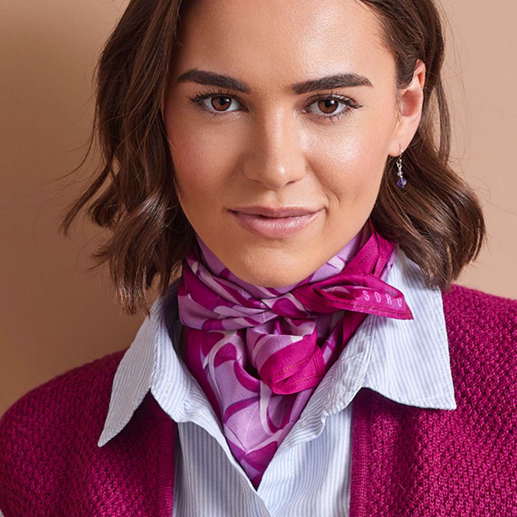 A close-up of a woman wearing a vibrant magenta and purple patterned silk scarf tied around her neck, styled over a white striped shirt and a magenta knit cardigan.