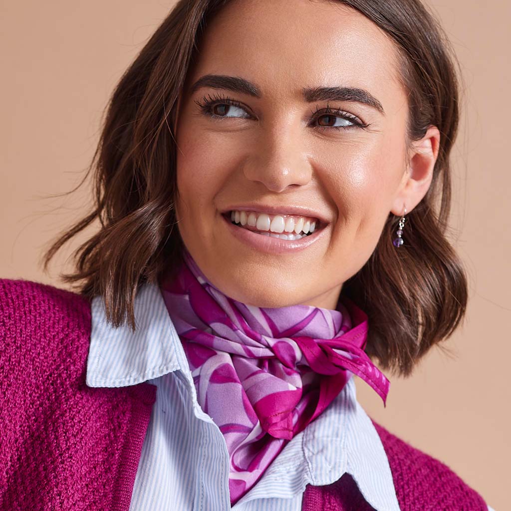 A smiling woman with brown shoulder-length hair wears a bright pink and purple patterned silk scarf tied around a white collared shirt, paired with a magenta knit cardigan. She is wearing a small dangling earring against a solid beige background.
