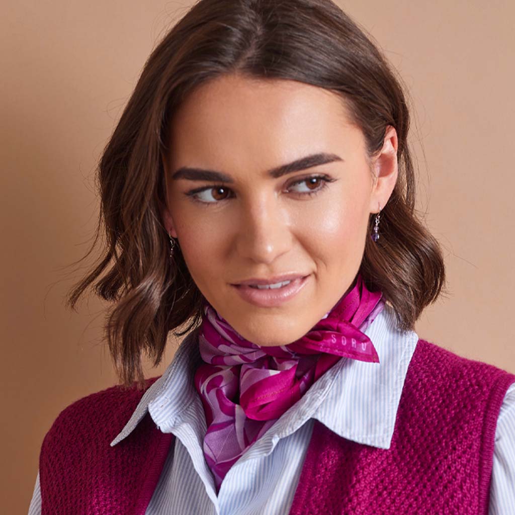 A woman with brown hair wearing a bright magenta and purple patterned neck scarf, a white and blue striped button-down shirt, and a textured magenta knit vest, set against a solid warm beige background.