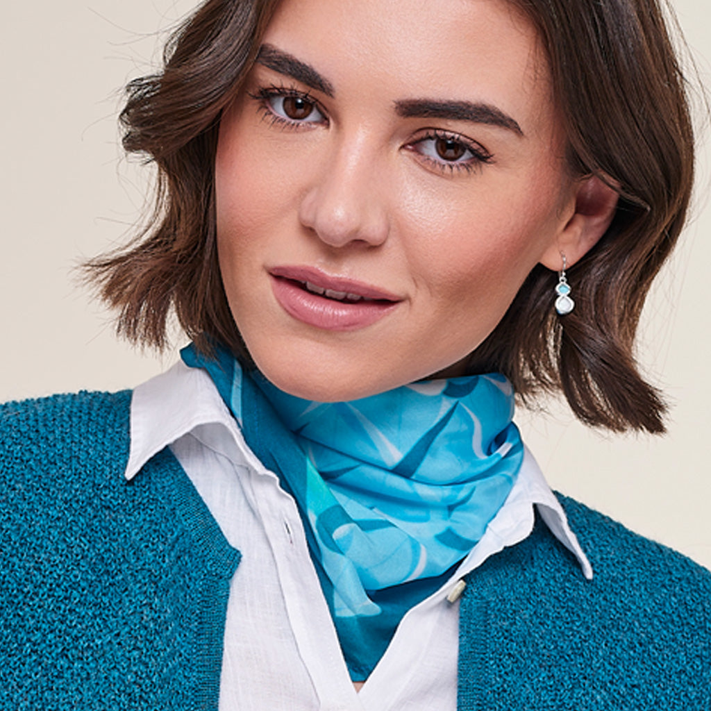 A close-up of a woman wearing a bright blue patterned silk scarf tied around her neck, a white collared shirt, and a teal knit cardigan. She is also wearing a small silver and white teardrop earring.