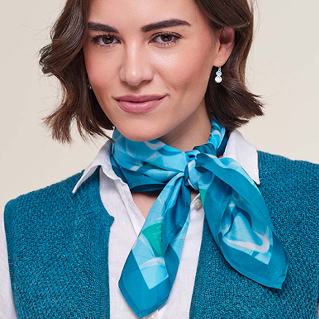 A woman wearing a bright blue and teal patterned silk scarf tied around her neck, styled with a white collared shirt and a teal knit vest against a light beige background.
