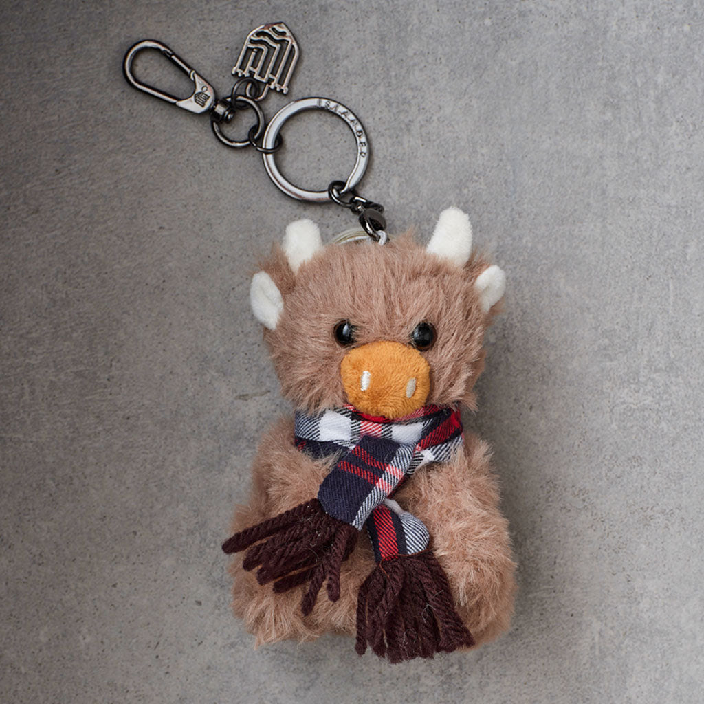 A plush toy keychain shaped like a fluffy brown animal with white horns, a plaid scarf, and silver metal keyring hardware, photographed on a gray textured background.