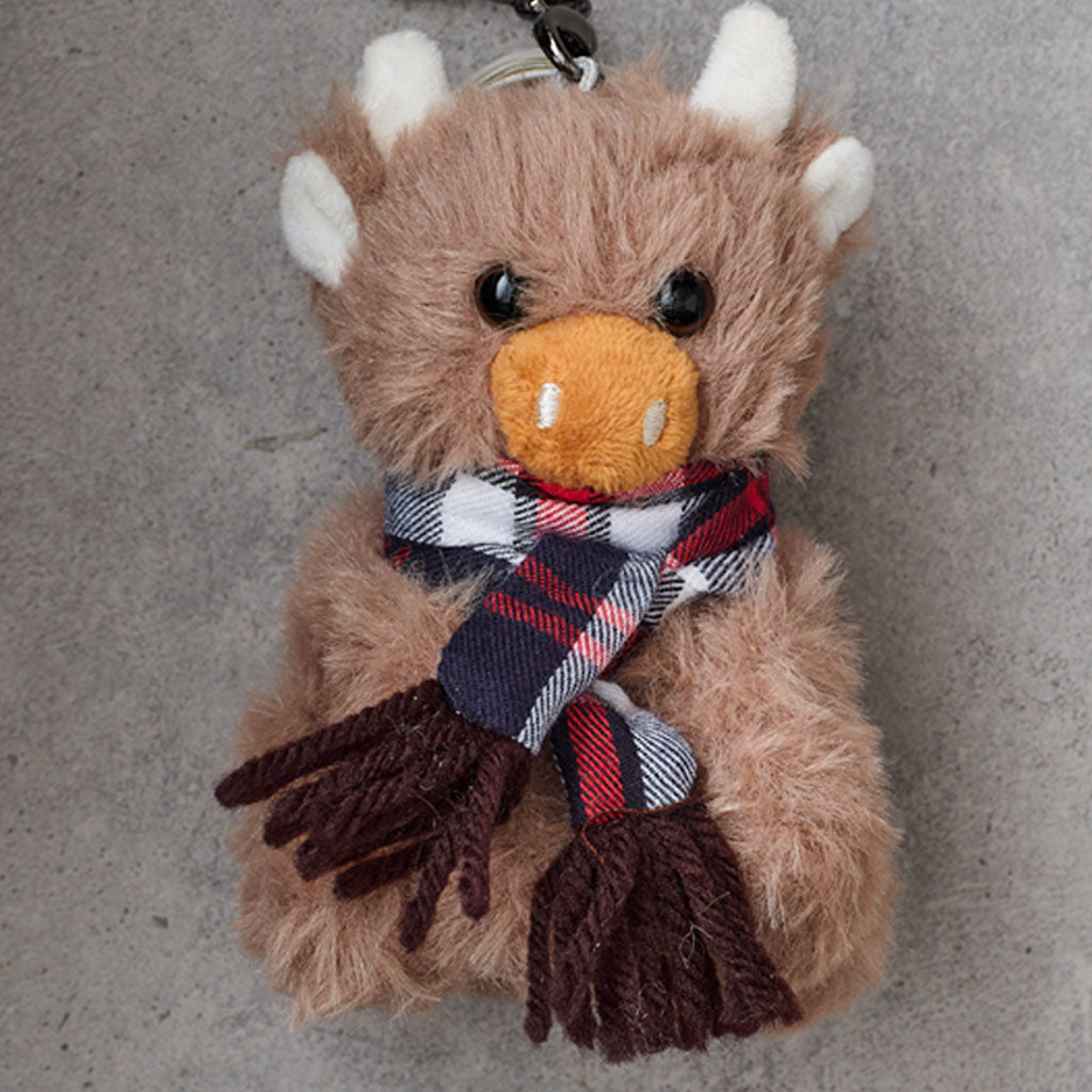 A plush toy keychain shaped like a fluffy brown animal with white horns, a stitched orange nose, and a plaid scarf in navy, red, white, and gray, sitting on a gray textured surface.