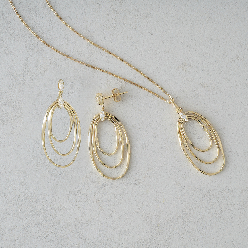 Gold-toned jewelry set featuring a delicate chain necklace and matching drop earrings, each with three nested oval loops, displayed on a light gray textured surface.