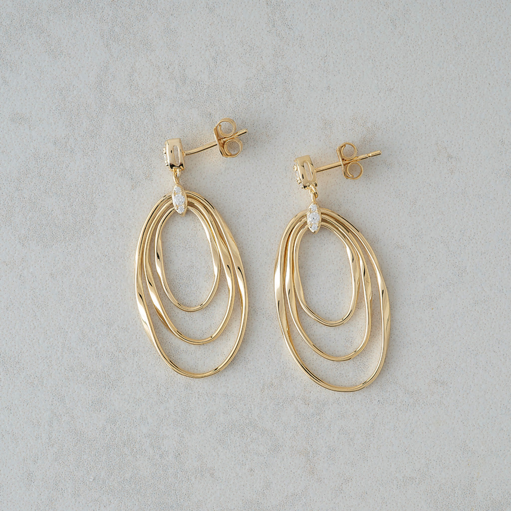 A pair of gold dangle earrings with multiple nested oval rings and a small clear stone accent, displayed on a light gray textured background.
