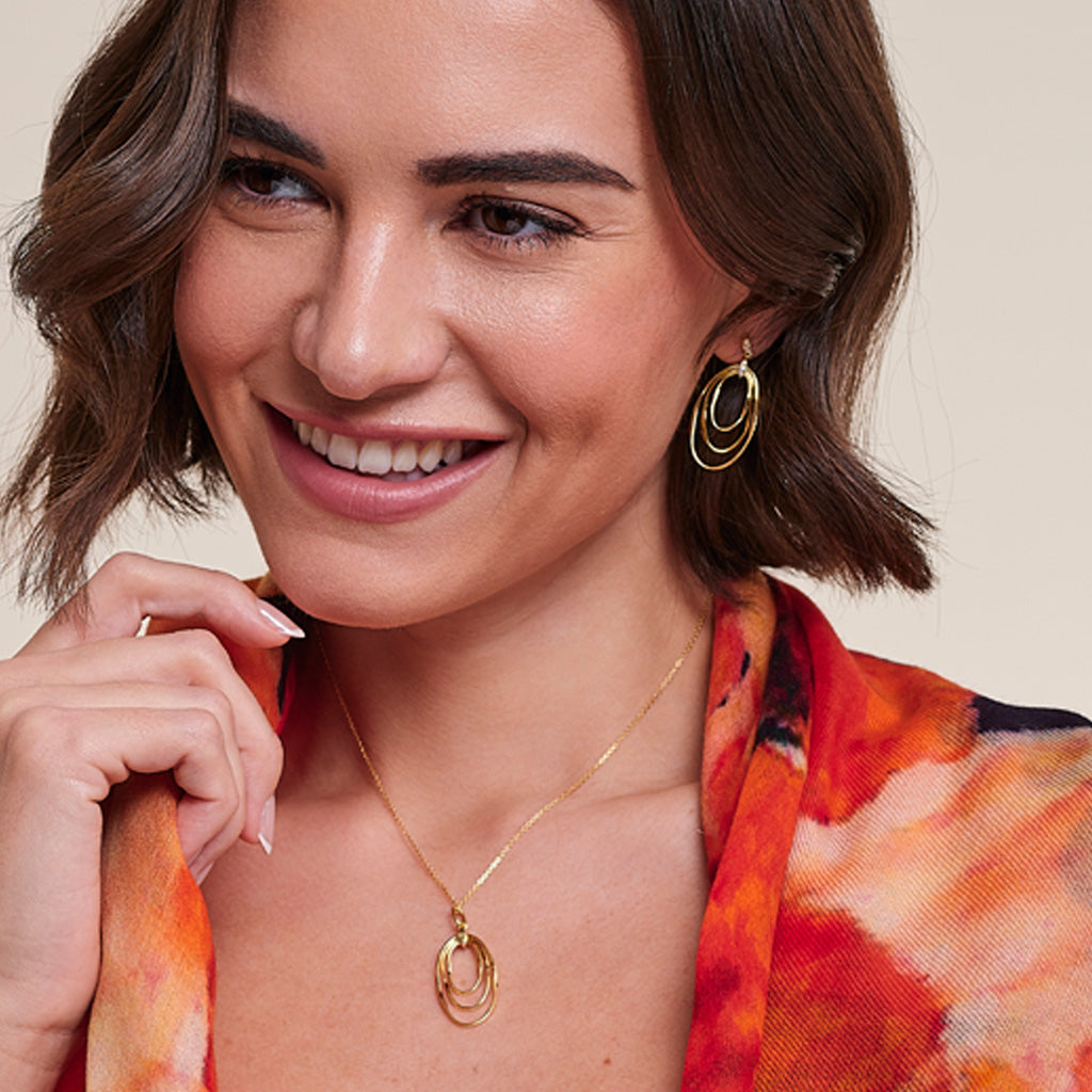 A smiling woman with short brown hair models a gold necklace and matching gold hoop earrings against a light beige background.
