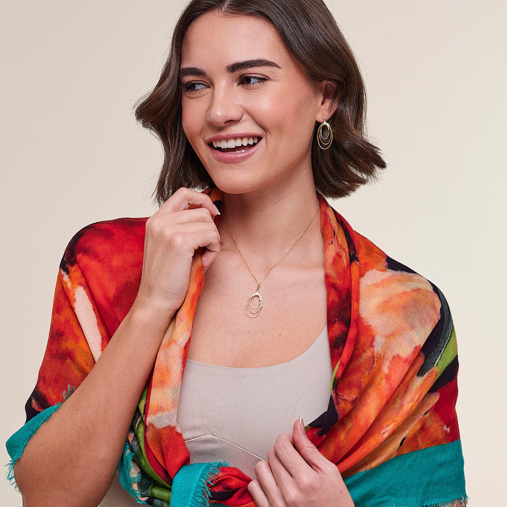 A woman smiling and wearing a vibrant multicolor abstract print scarf draped over her shoulders, paired with a beige tank top and gold jewelry, against a solid light beige background.