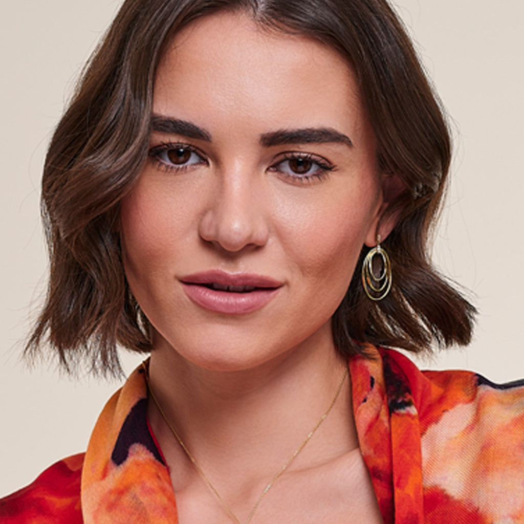 A close-up portrait of a woman with short brown hair wearing gold double-ring hoop earrings and a vibrant orange, red, and black patterned top against a light beige background.
