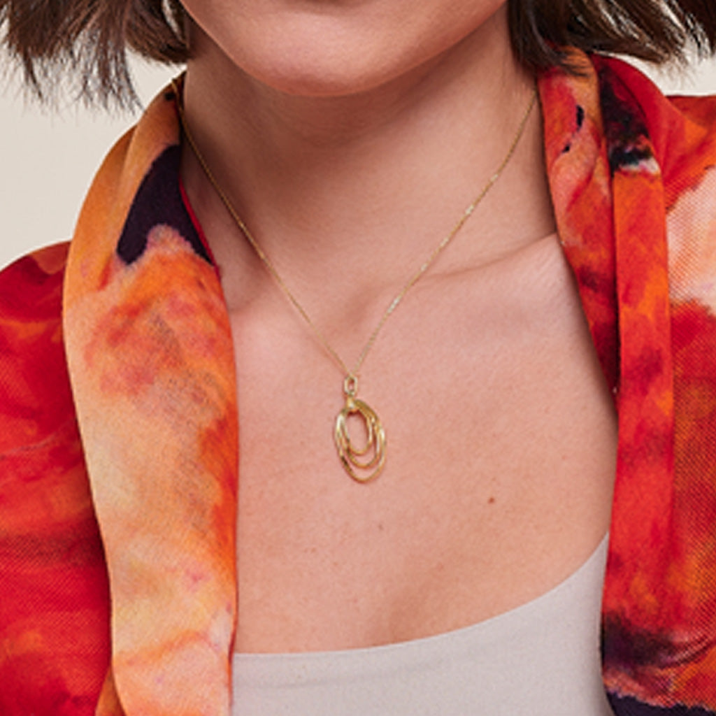 A close-up of a model wearing a delicate gold necklace with a double-ring pendant, styled with a vibrant orange and red patterned scarf.