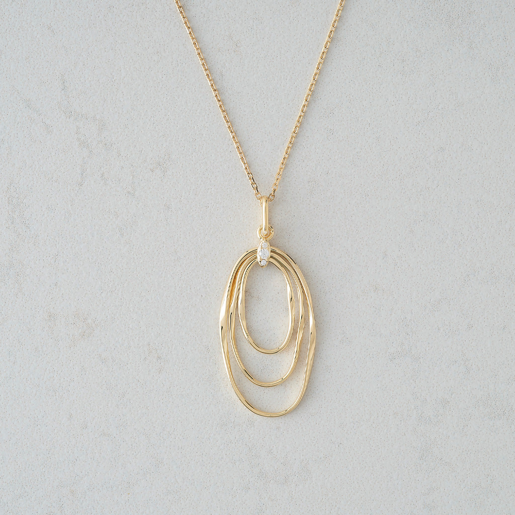 A delicate gold chain necklace with a multi-layered, organic-shaped oval pendant featuring a small clear crystal accent at the top, displayed on a light gray textured background.