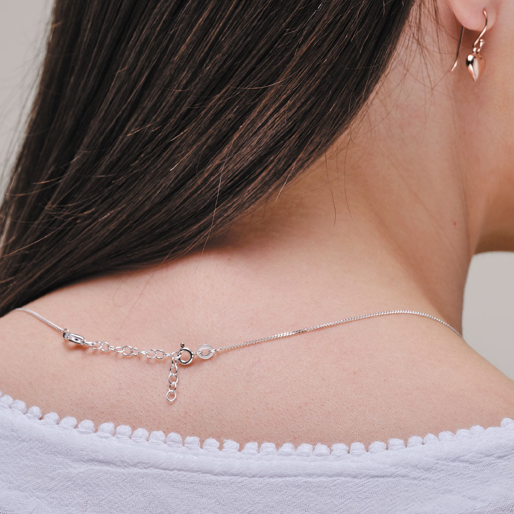 A close-up lifestyle shot of a model's neck and upper back wearing a delicate silver chain necklace with a small charm and a matching silver hoop earring.
