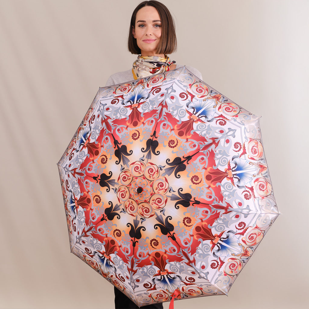 A woman holding up a large open umbrella to display its intricate pattern. The umbrella features a symmetrical mandala-style design with red, orange, black, and blue motifs on a white background.