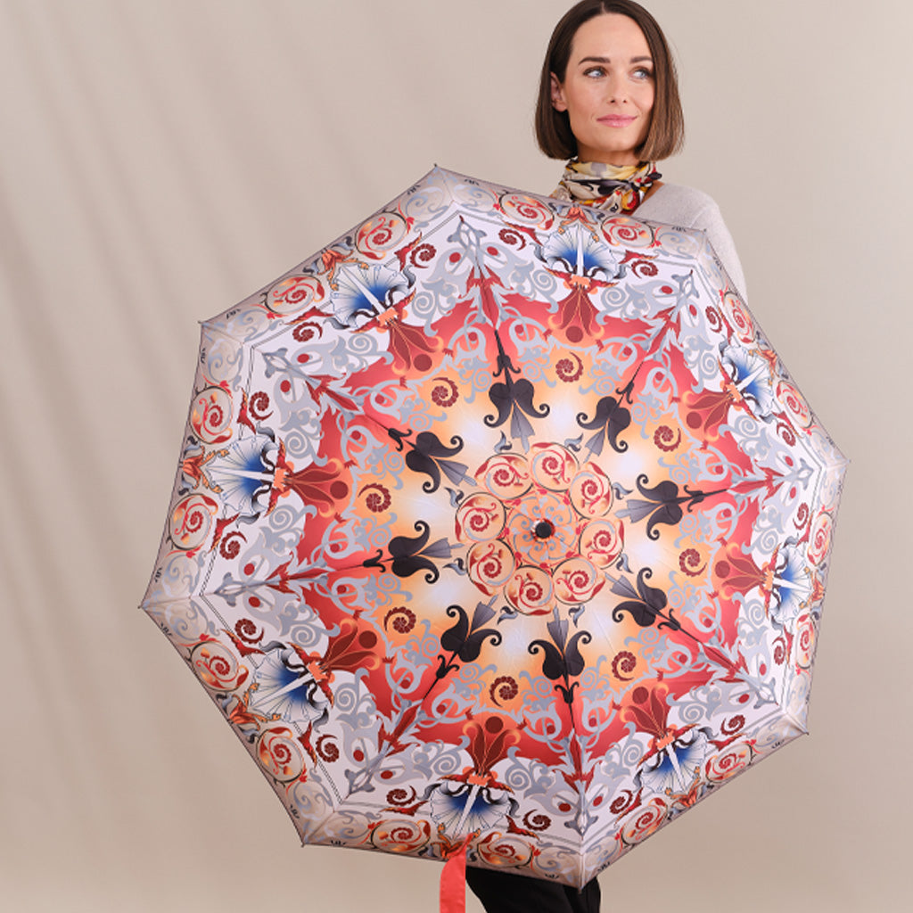 A woman holding a large, open umbrella with an intricate, symmetrical mandala-style pattern in red, orange, black, and blue on a light background. She is wearing a gray top and a patterned scarf, set against a plain beige studio backdrop.
