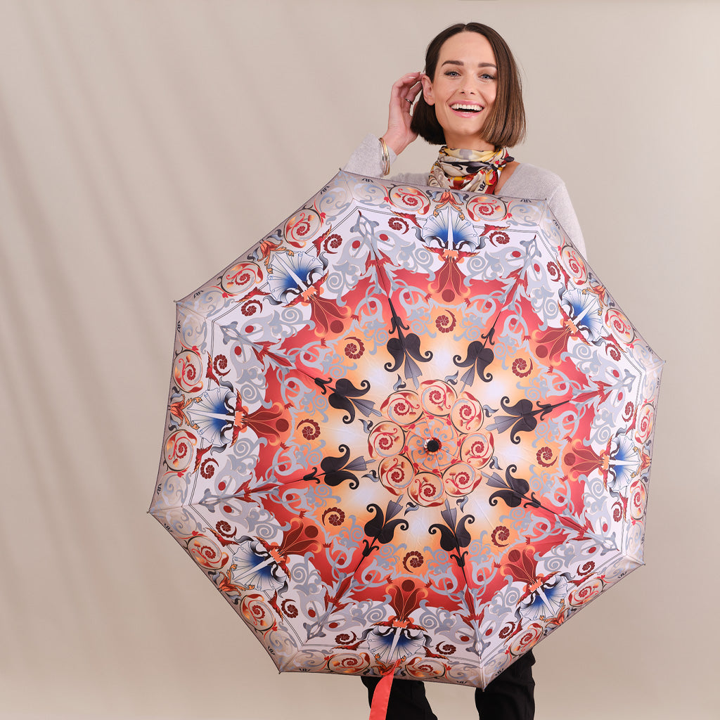 A woman holding a large, open umbrella with an intricate, symmetrical mandala-style pattern in red, orange, black, and blue on a light background. She is wearing a gray top and a patterned scarf, set against a plain beige studio backdrop.