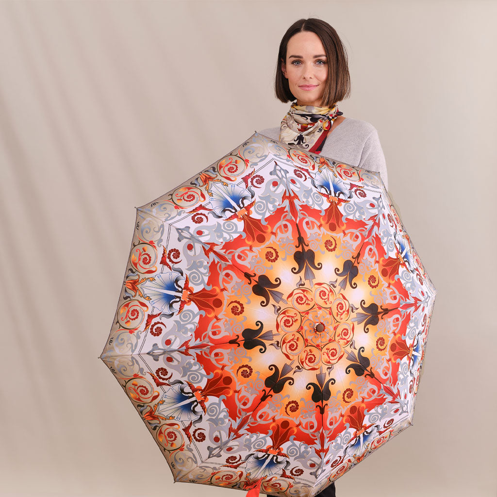 A woman holding a large, open umbrella with an intricate, symmetrical mandala-style pattern in red, orange, black, and blue on a light background. She is wearing a gray top and a patterned scarf, set against a plain beige studio backdrop.