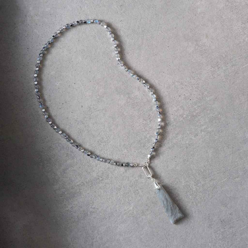 A beaded necklace with small clear faceted beads and a light gray stone pendant, laid on a textured gray surface.
