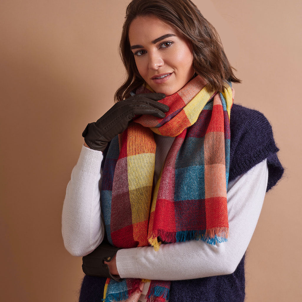 A woman wearing a multicolor plaid scarf with red, yellow, blue, and orange blocks, styled with a navy knit sweater vest, white long-sleeve shirt, and brown leather gloves against a solid beige background.