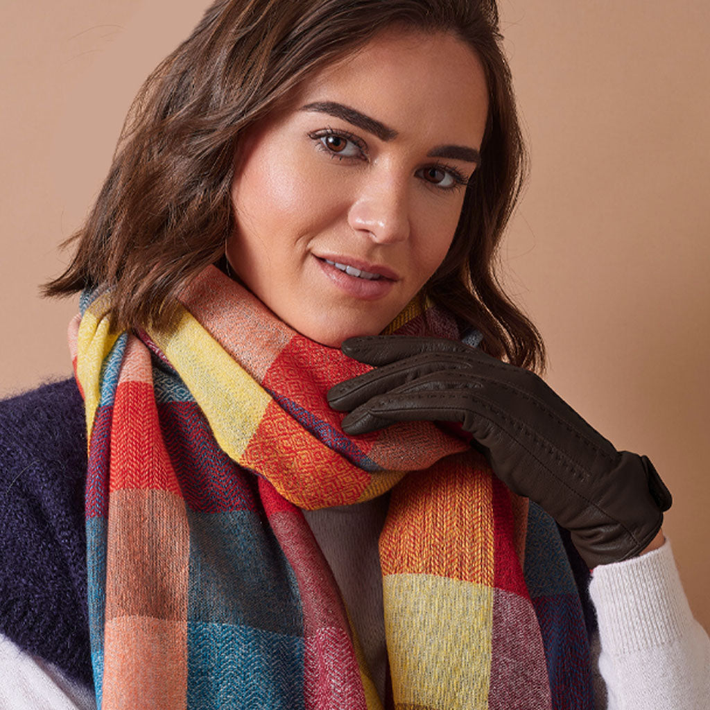 A woman wearing a thick multicolor plaid scarf in shades of red, yellow, blue, and orange, paired with a dark brown glove and a navy knit sweater, set against a solid beige background.