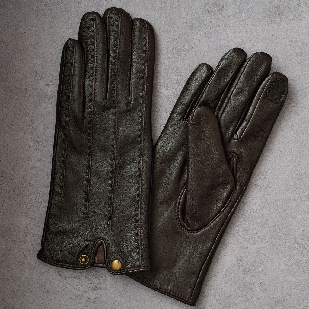 A pair of dark brown leather gloves, one showing the back with decorative stitching and snap closures, and the other showing the palm with a touch patch on the fingertip, laid flat on a textured gray background.
