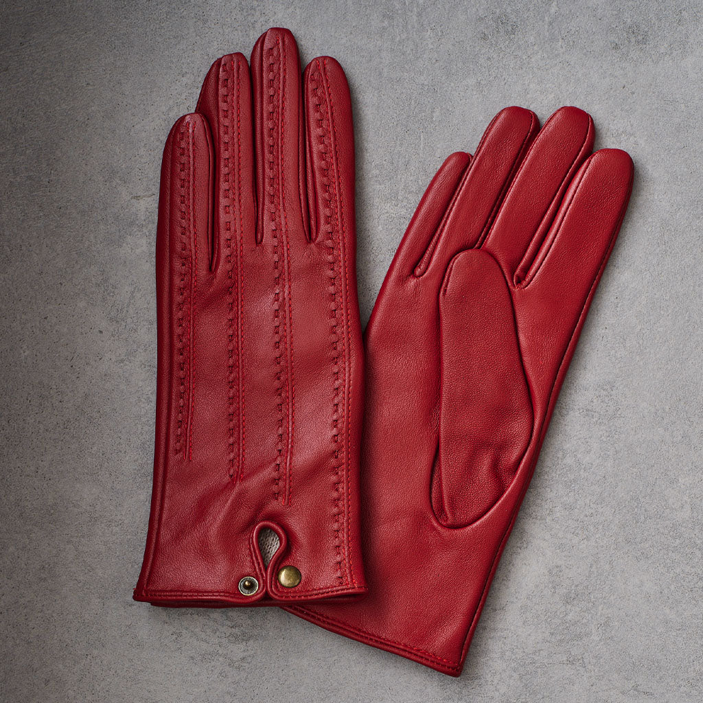 A pair of red leather gloves laid flat on a textured gray background, showing the back of one glove with decorative stitching and a small gold-tone snap closure, and the palm side of the other glove.