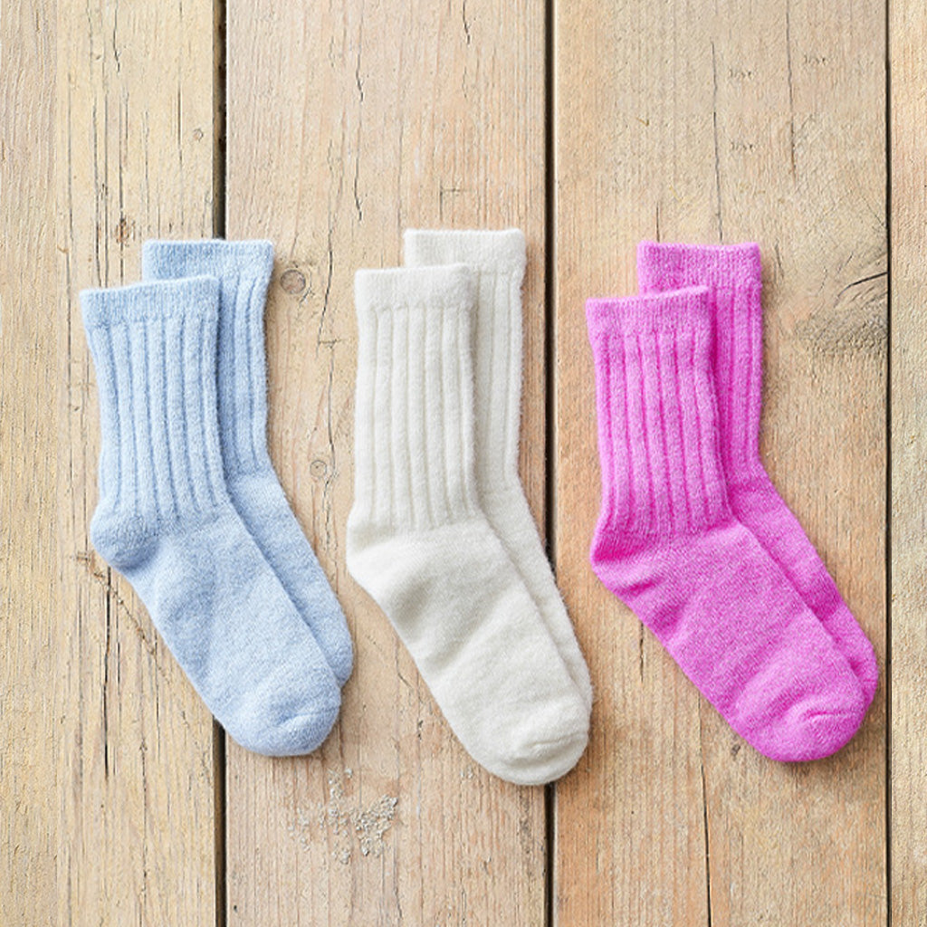 Three pairs of ribbed crew socks in light blue, off-white, and bright pink, arranged side by side on a light brown wooden plank background.