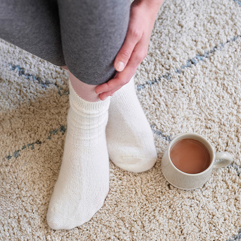 A person wearing cream-colored knit socks sits on a beige carpet next to a ceramic mug of coffee.