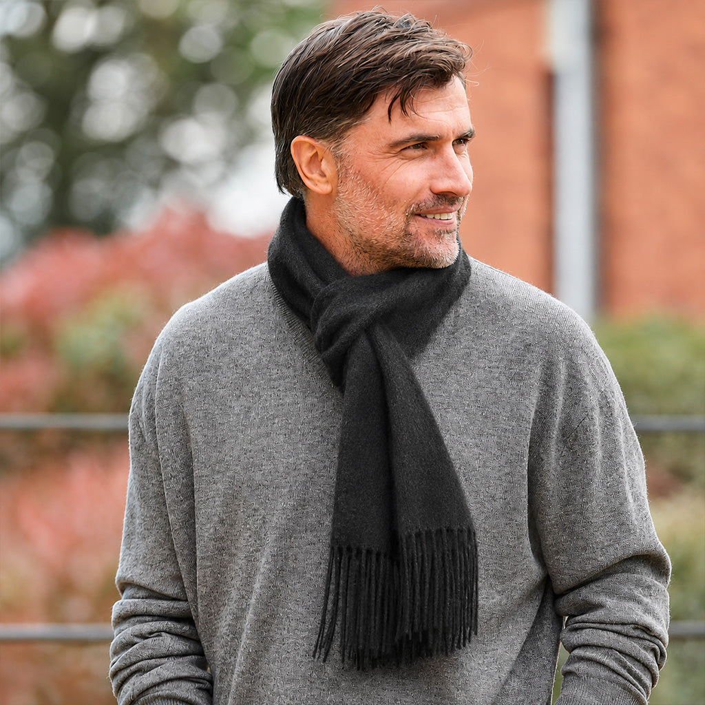 A man wearing a thick black knit scarf and a gray long-sleeve sweater, photographed outdoors with a blurred brick wall and foliage in the background.