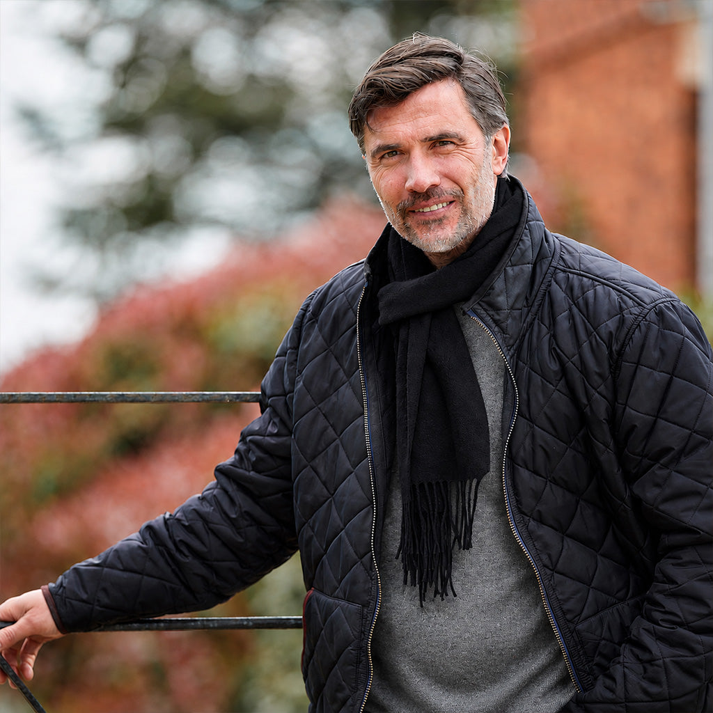 A man wearing a black quilted jacket and a black scarf, standing outdoors with a blurred background of trees and a brick wall.