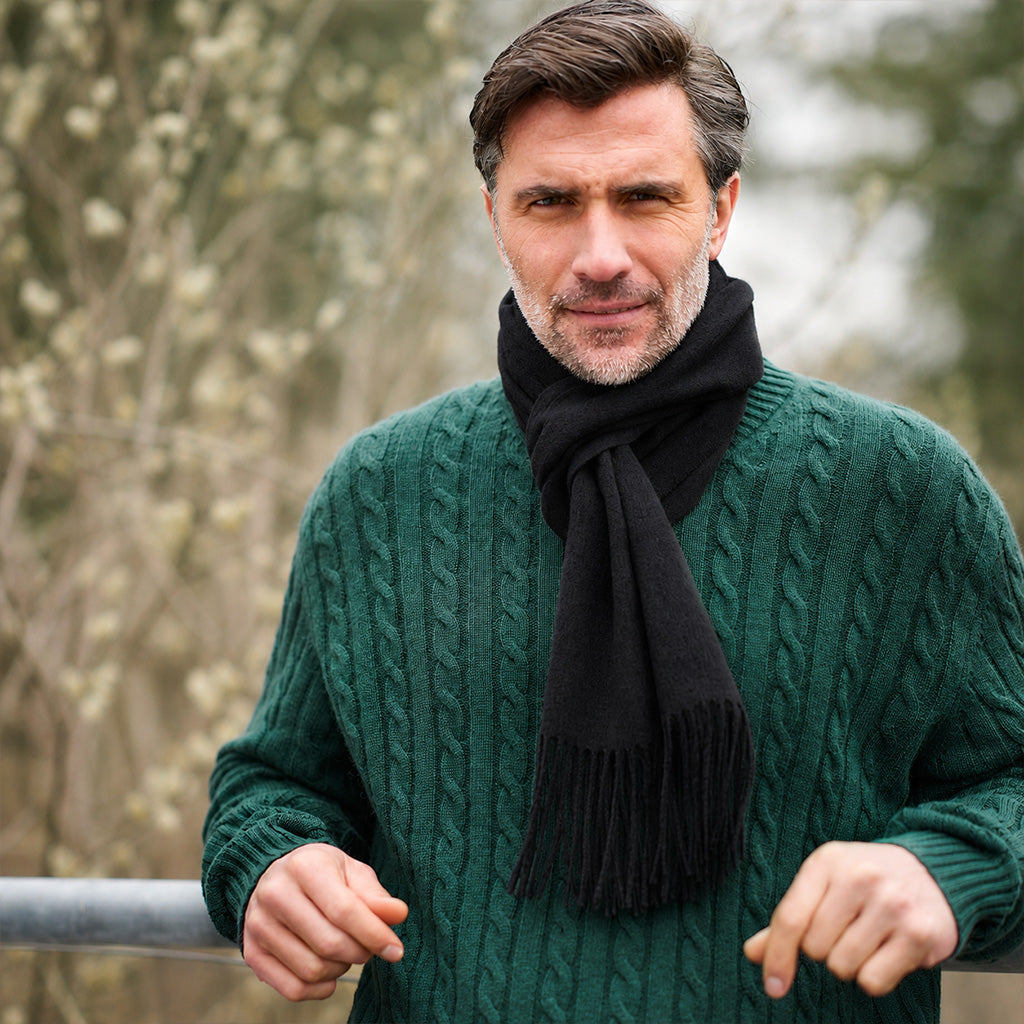 A man wearing a dark green cable-knit sweater and a black knit scarf, standing outdoors with a blurred natural background.