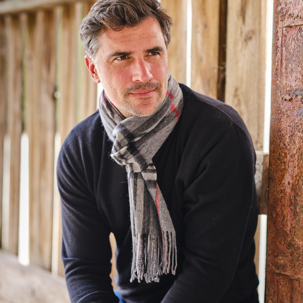 A man wearing a dark navy sweater and a gray plaid scarf with red and black stripes, leaning against a rustic wooden wall.