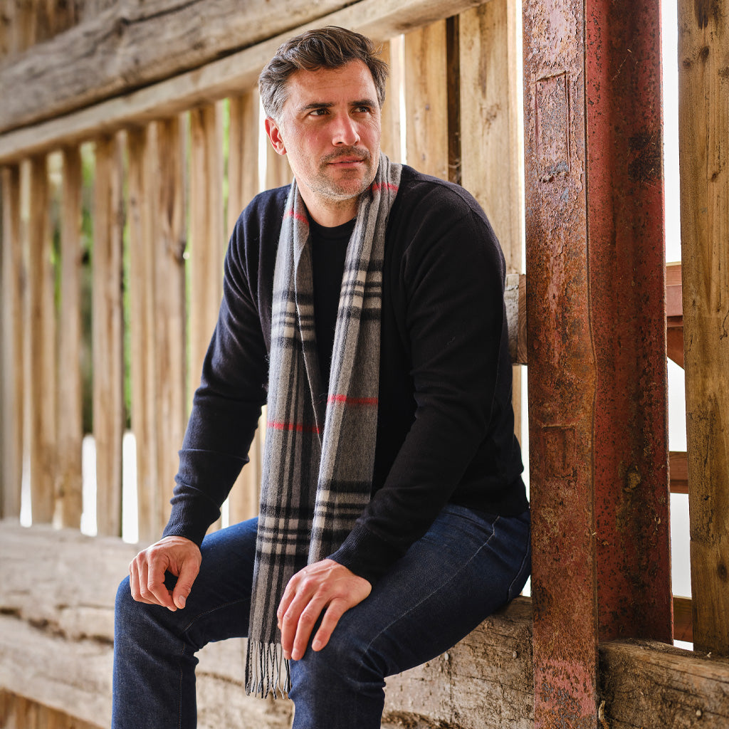 A man wearing a black long-sleeve shirt, blue jeans, and a gray plaid scarf with red and white stripes, sitting on rustic wooden steps.