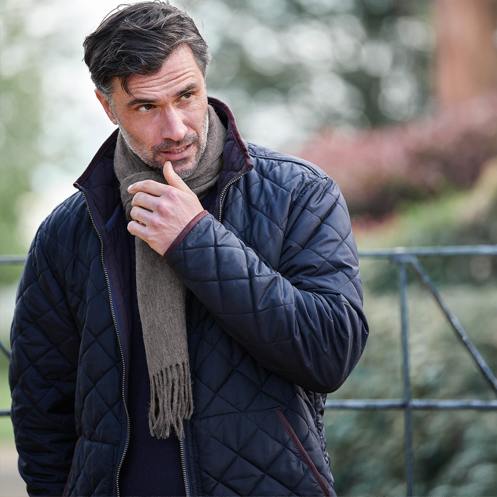 A man wearing a dark navy quilted jacket and a brownish-gray knit scarf, standing outdoors with a blurred background.