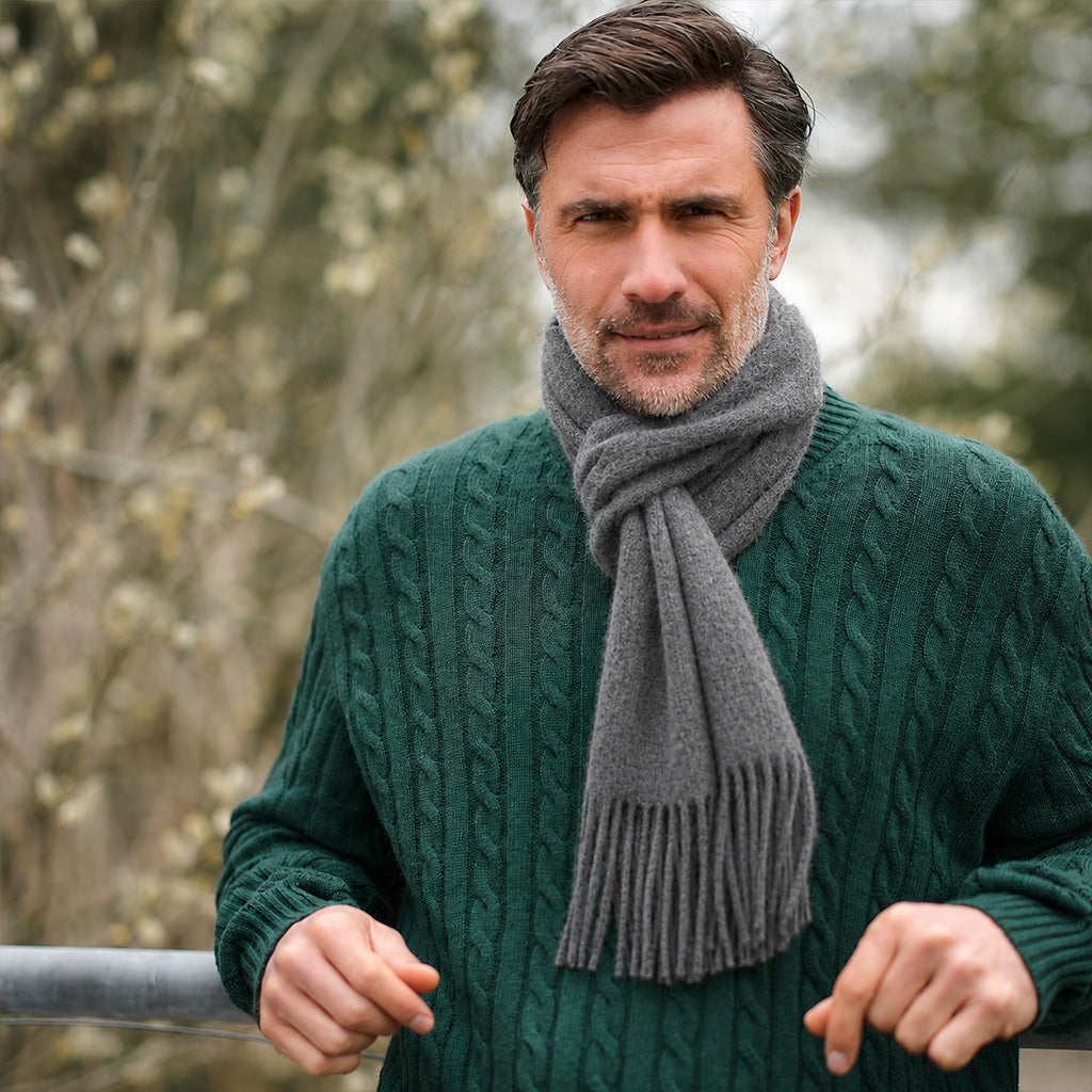 A man wearing a dark green cable-knit sweater and a gray knit scarf, standing outdoors with a blurred natural background.