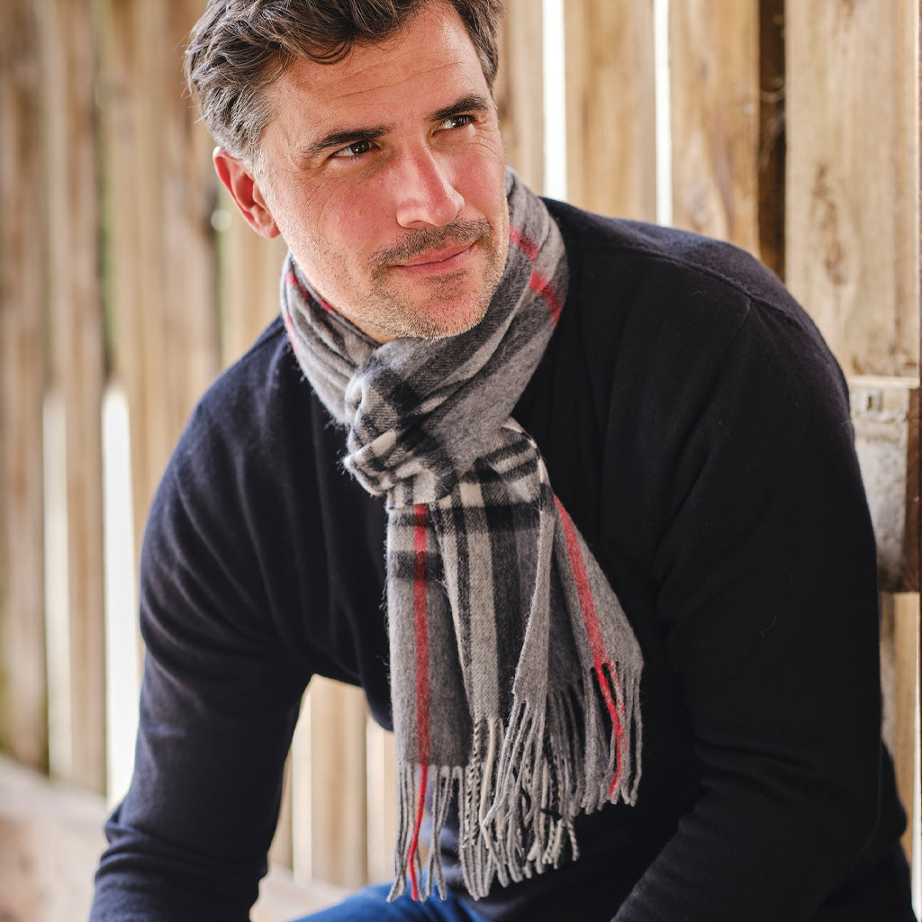 A man wearing a dark navy sweater and a gray plaid scarf with red and white stripes, posed against a rustic wooden background.