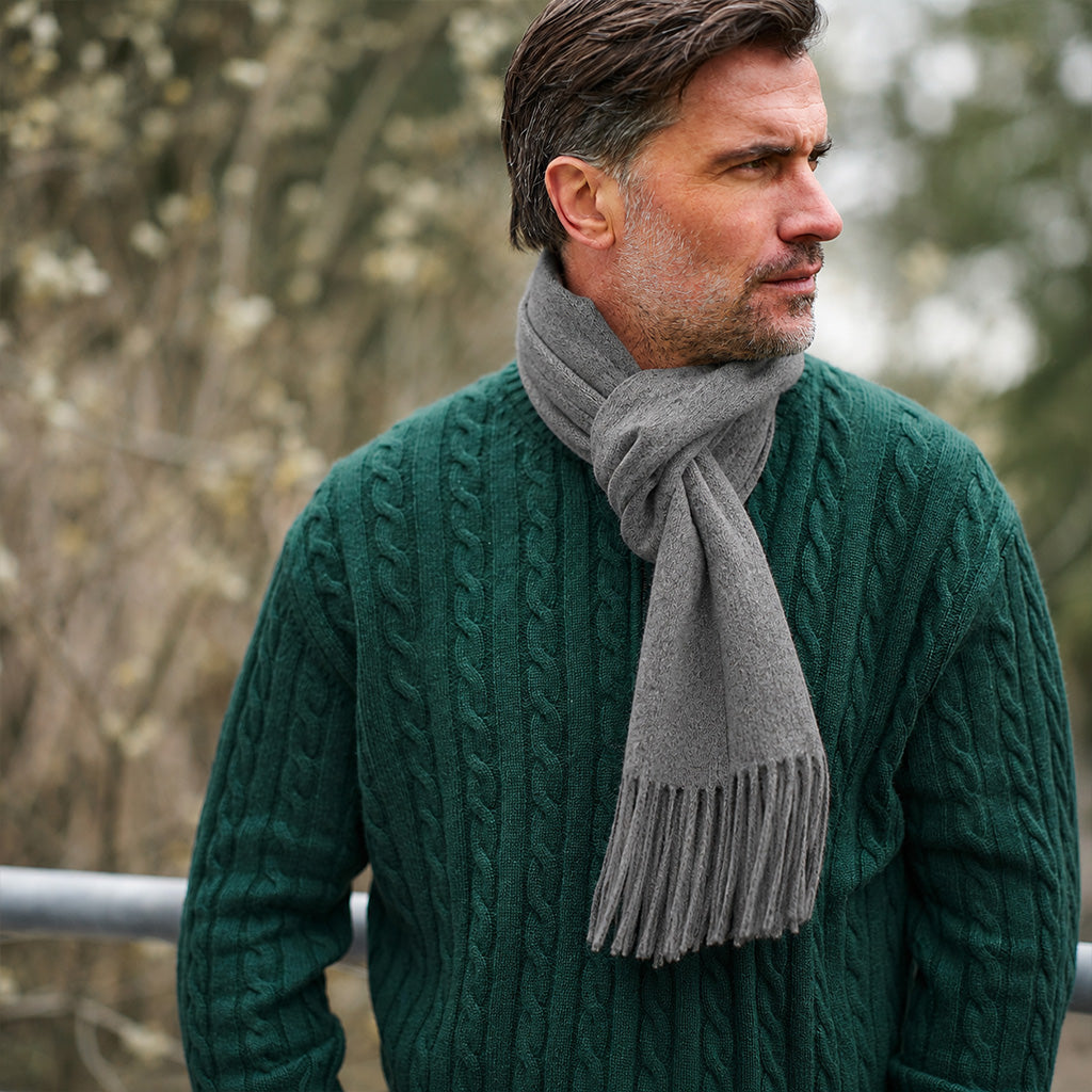 A man wearing a dark green cable-knit sweater and a light gray knit scarf, photographed outdoors with a blurred natural background.