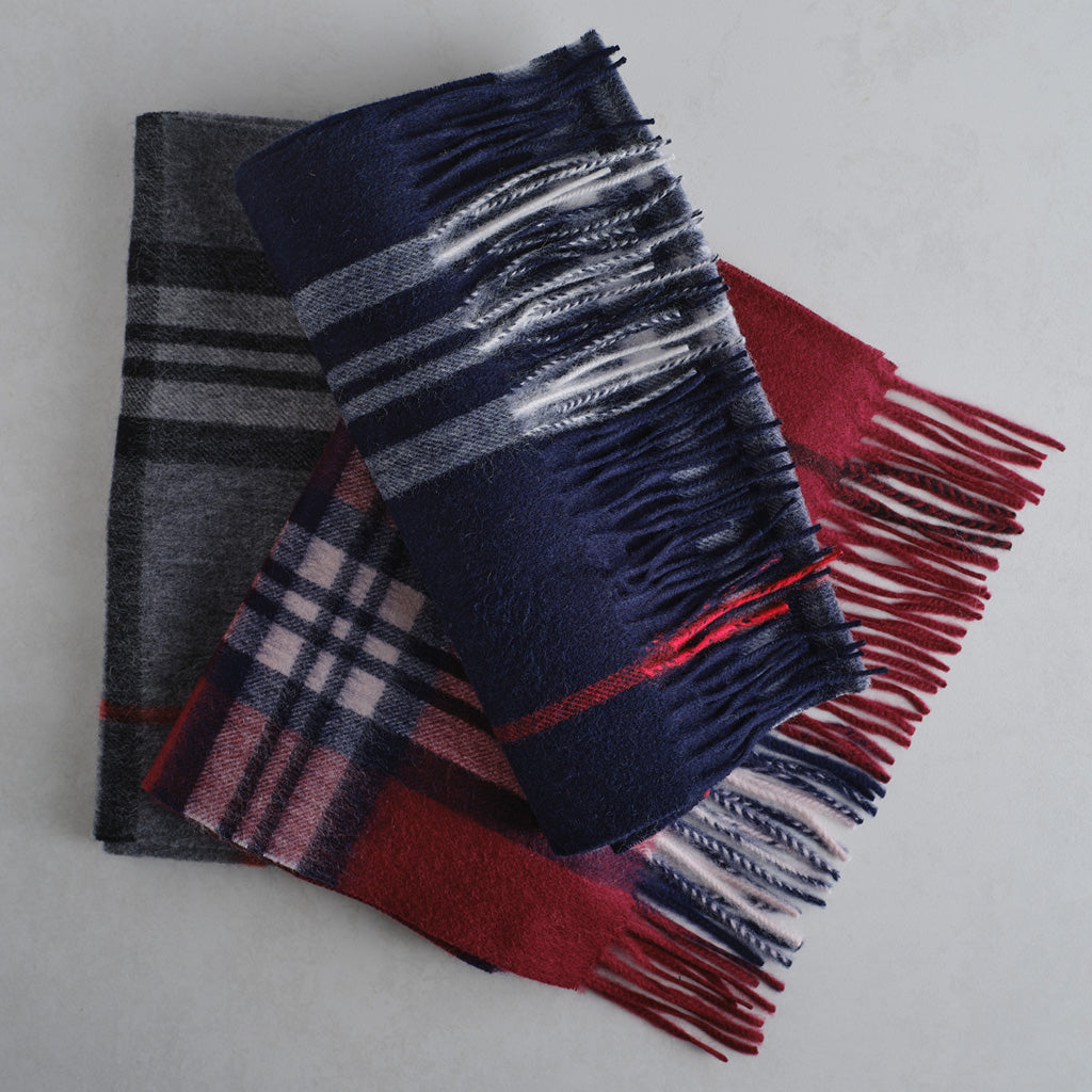 Three folded plaid and striped wool scarves with fringed edges, stacked on a light gray surface. The top scarf is navy blue with white and gray stripes, the middle scarf is red and pink plaid, and the bottom scarf is dark gray and black plaid.