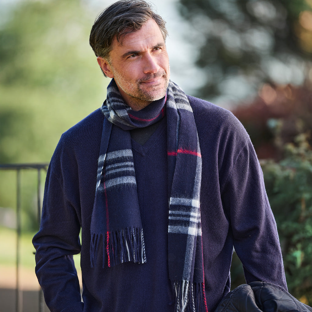 A man wearing a navy blue turtleneck sweater and a dark gray and white plaid scarf with thin red stripes, standing outdoors with a blurred green background.