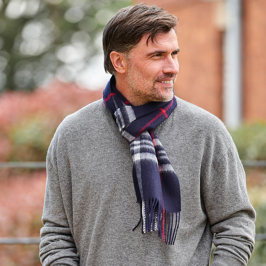 A man wearing a gray knit sweater and a navy blue plaid scarf with red and white stripes, standing outdoors with a blurred brick wall and greenery in the background.
