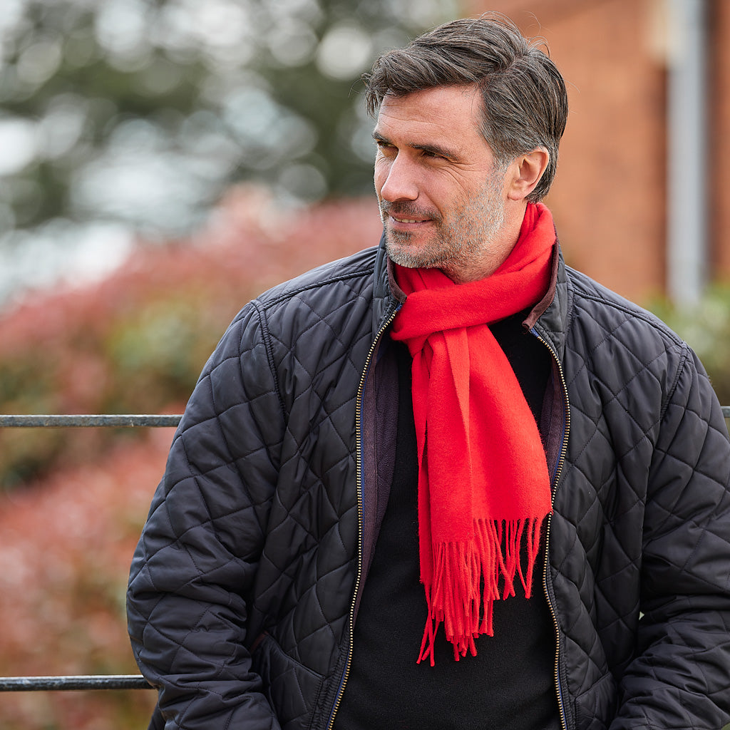 A man wearing a bright red fringed scarf and a black quilted jacket, photographed outdoors with a blurred background.